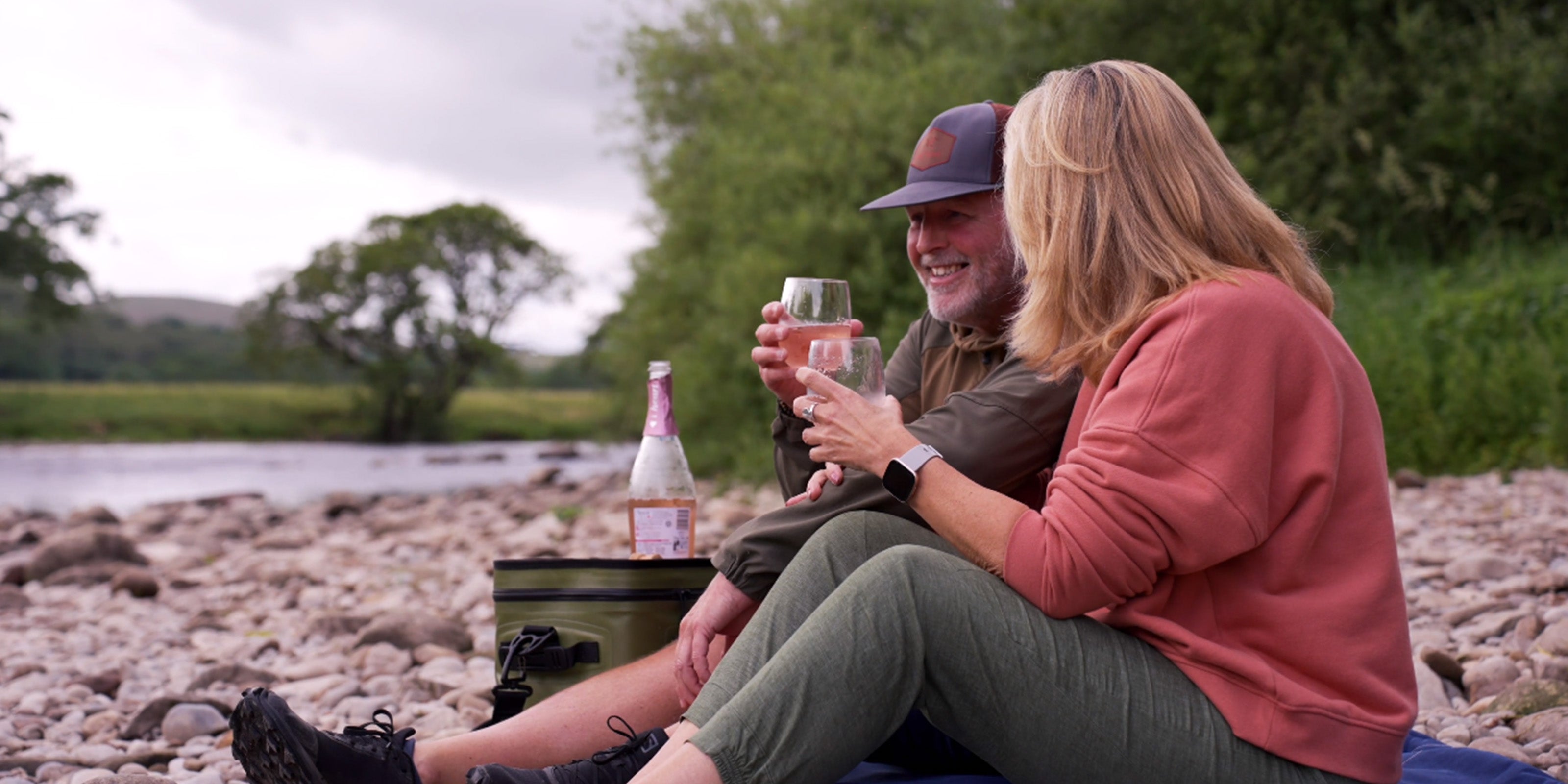 Image of a man and woman on a pebbled beach having a picnic next to a green Softcool cooling bag