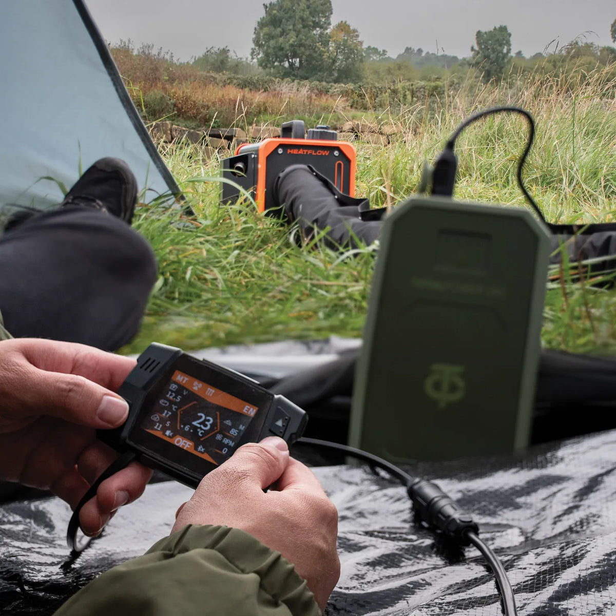 A person outdoors in a tent in a field uses a controller connected to a Heatflow diesel heater sitting in the background near a tent, with trees and an overcast sky visible. 