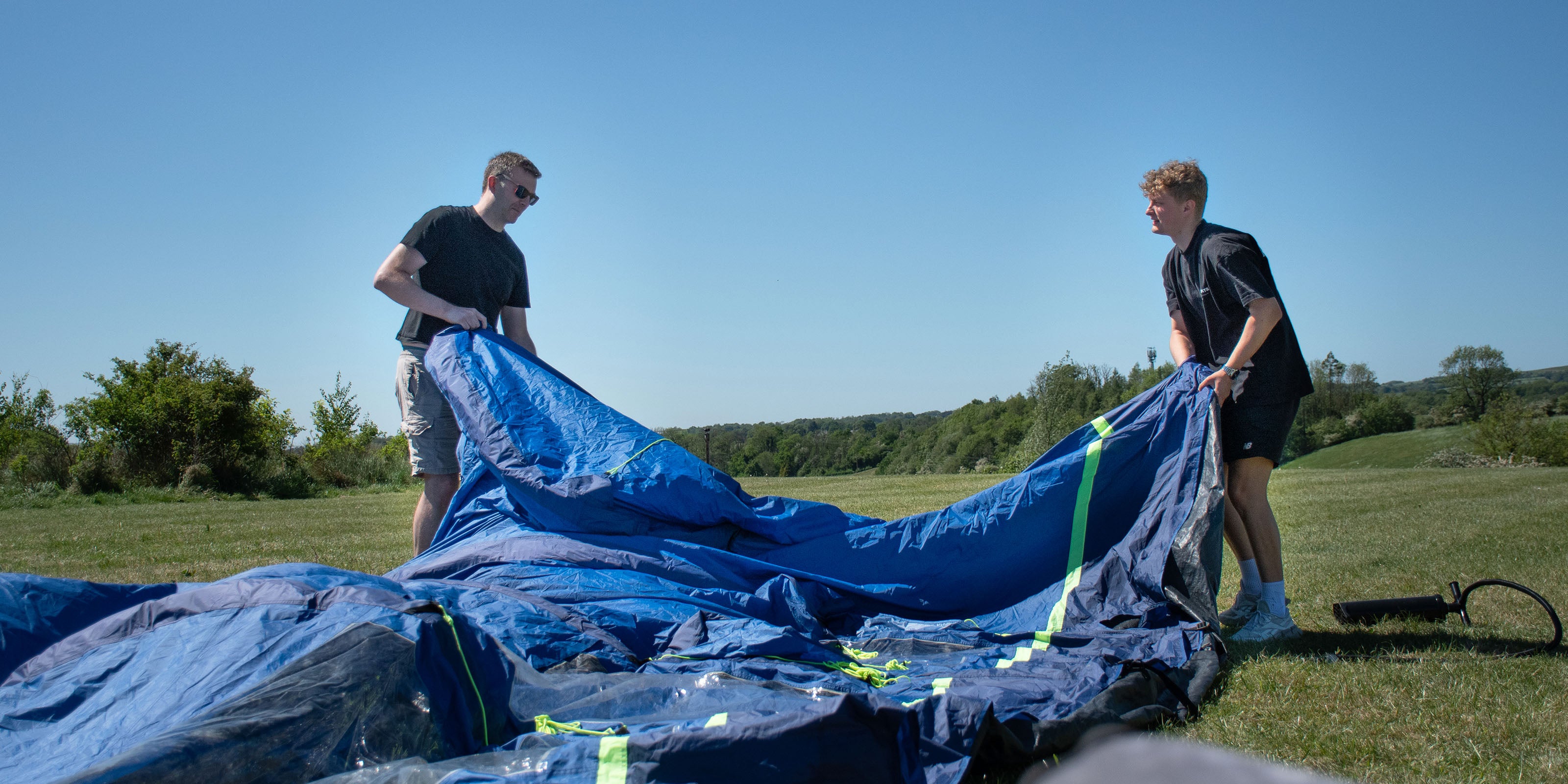 Two people working with a large blue tent in an open field on a clear day.